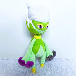 May include: A plush doll of a green creature with a white head, purple eyes, and yellow accents. The doll has a smiling expression and is made of soft fabric. The creature's hands and feet are a darker color.