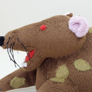 K&ouml;nnte beinhalten: Ein braunes Pl&uuml;schrattenspielzeug mit einem rosa Ohr, gr&uuml;nen Flecken und einem roten Mund. Die Ratte hat schwarze Schnurrhaare und eine angen&auml;hte rote Nase.
