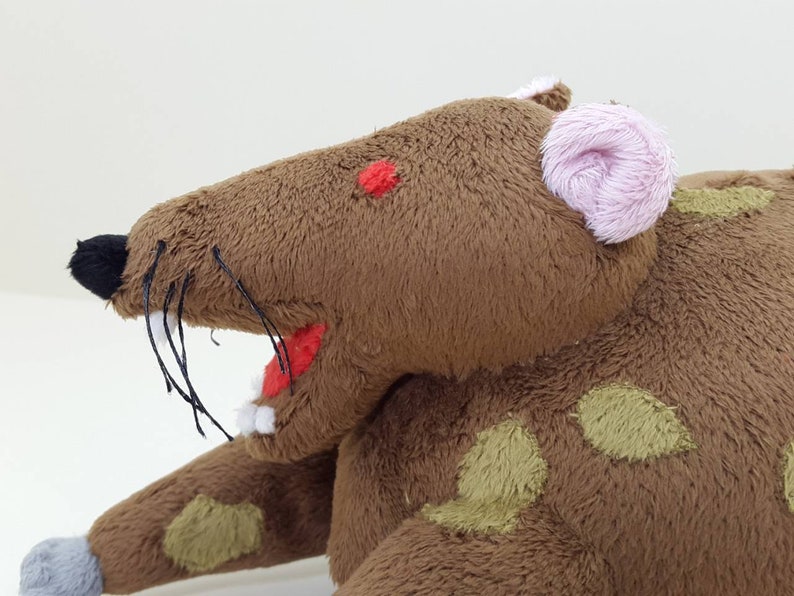 K&ouml;nnte beinhalten: Ein braunes Pl&uuml;schrattenspielzeug mit einem rosa Ohr, gr&uuml;nen Flecken und einem roten Mund. Die Ratte hat schwarze Schnurrhaare und eine angen&auml;hte rote Nase.