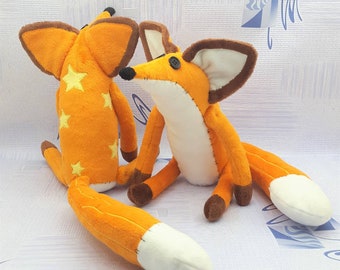 May include: Two plush orange fox toys with brown accents and white-tipped tails. One fox has yellow star appliqués. The foxes have button eyes and are made of soft fabric. The toys are designed to look like a cartoon fox.