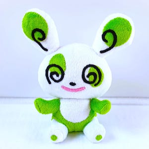 Könnte beinhalten: Ein weißes Plüschkaninchen mit grünen Akzenten. Das Kaninchen hat grüne Ohren, Pfoten und grüne Flecken auf den Wangen. Schwarze Wirbelmuster sind um die Augen gestickt. Es hat ein rosa gesticktes Lächeln.