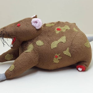 K&ouml;nnte beinhalten: Ein braunes Pl&uuml;schrattenspielzeug mit roten Augen, einer rosa Nase und gr&uuml;nen und roten Flecken. Die Ratte liegt auf dem Bauch mit ausgestreckten Beinen.