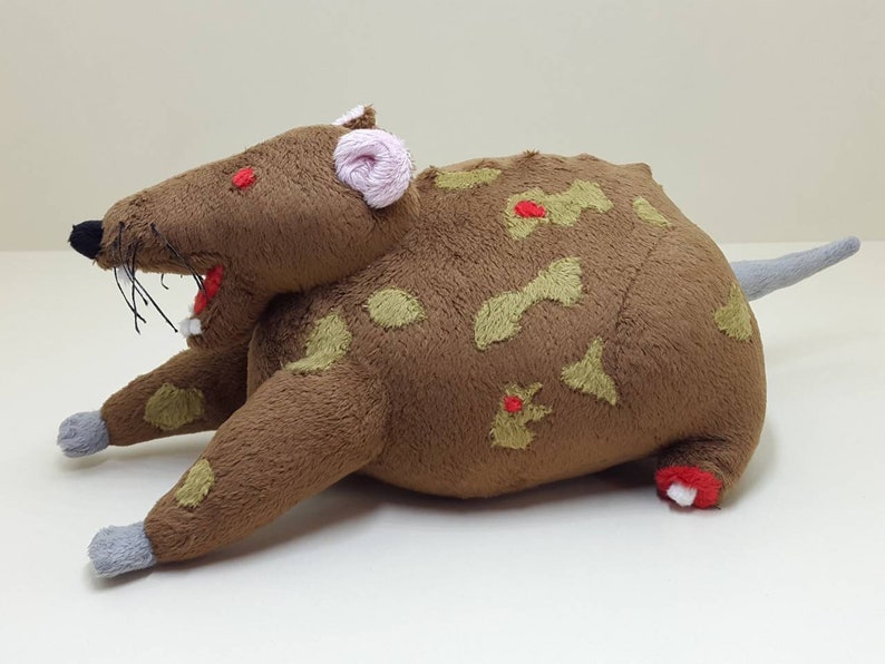 K&ouml;nnte beinhalten: Ein braunes Pl&uuml;schrattenspielzeug mit roten Augen, einer rosa Nase und gr&uuml;nen und roten Flecken. Die Ratte liegt auf dem Bauch mit ausgestreckten Beinen.