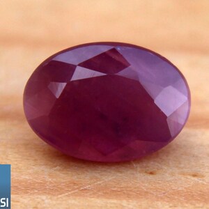 Peut inclure: Une pierre précieuse rouge foncé, taillée en forme ovale et à facettes. La gemme est posée sur une surface en bois brun clair. Le logo GSI est dans le coin inférieur gauche.
