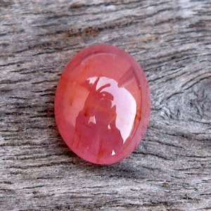 May include: A polished, oval-shaped gemstone with a glossy, reflective surface. The gem has a reddish-pink hue with variations in color, set against a textured, gray wooden background. The stone's smooth surface reflects light.