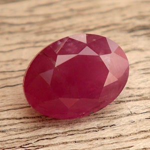 Peut inclure: Une pierre précieuse ovale de couleur rouge foncé avec une taille facetée. La pierre précieuse est posée sur un fond en bois brun clair.