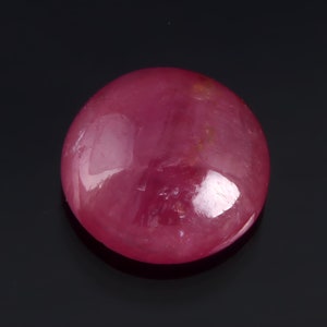 Könnte beinhalten: Ein runder, cabochon geschliffener Rubin mit einer tief roten Farbe. Der Stein ist poliert und hat eine glänzende Oberfläche.