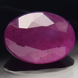 May include: A vibrant pink oval-shaped gemstone with a faceted cut. The gemstone has a slightly cloudy appearance with small black inclusions.