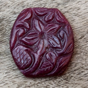 Puede incluir: Una piedra preciosa de rubí tallada con un diseño floral. El rubí es de color rojo intenso y tiene un alto pulido.