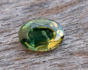 May include: An oval-cut gemstone with a gradient of colors, transitioning from a deep green to a golden yellow. The gem is faceted, reflecting light and set against a weathered, wooden surface.