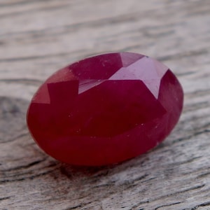 Può includere: Una pietra preziosa di colore rosso intenso con forma ovale sfaccettata. La pietra preziosa è appoggiata su una superficie di legno.