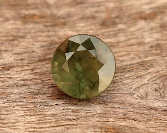 May include: A faceted, round gemstone with a pale green hue. The gem is set against a textured, brown wooden surface. The facets create a play of light and shadow, highlighting the gem's clarity.