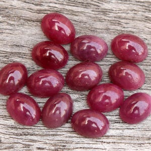 Könnte beinhalten: Eine Sammlung von 12 ovalen, polierten Rubin-Cabochons in verschiedenen Rosatönen und Rottönen. Die Steine sind auf einer Holzoberfläche angeordnet.