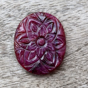 May include: A carved ruby gemstone featuring a floral design. The gemstone is oval shaped and has a deep red colour.