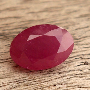 May include: A deep red oval-shaped gemstone with a faceted cut. The gemstone is sitting on a wooden surface.