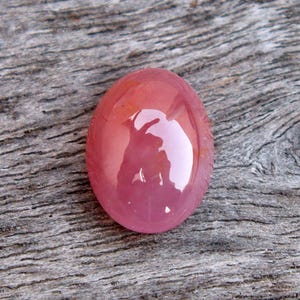 Puede incluir: Una piedra preciosa rosa lisa de forma ovalada con un acabado de alto brillo. La piedra preciosa se coloca sobre un fondo de madera.