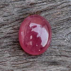 Op de afbeelding: Een gladde, ovale, roze edelsteen met een hoogglans afwerking. De edelsteen is geplaatst tegen een houten achtergrond.