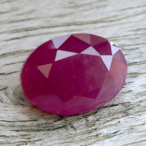 Puede incluir: Una gema de rubí facetada, de forma ovalada. La gema es de un color rojo intenso y rico con múltiples facetas que capturan la luz. La gema está sobre una superficie de madera clara y envejecida.