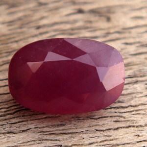 May include: A deep burgundy, oval-cut gemstone with multiple facets. The gem's surface reflects light, showcasing its cut. The stone is set against a rustic wooden background, highlighting its colour and cut.