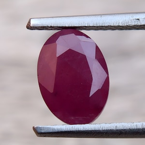Puede incluir: Una piedra preciosa ovalada de color rojo oscuro con un corte facetado. La piedra preciosa está sujeta por unas pinzas de metal.