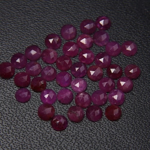 May include: A collection of ruby red faceted gemstones, each with a round shape and a polished finish. The stones are arranged in a loose pile on a dark gray surface.