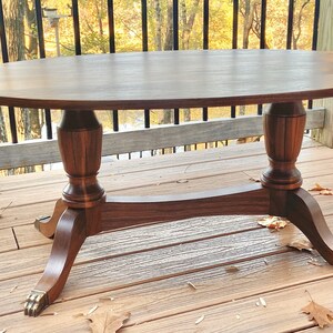 May include: A dark wood oval coffee table with a simple design. The table has a turned pedestal base with a decorative band and sits on four legs with brass paw feet.