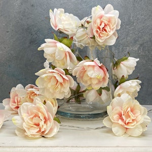 May include: Artificial peony flowers in cream and peach hues, some in a clear glass vase, others scattered on a white surface. A string of fairy lights is wrapped around the flowers, creating a soft glow.