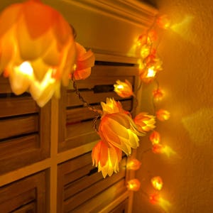 Op de afbeelding: Een decoratieve lichtslinger met bloemvormige lampjes in geel en oranje. De lampjes geven een warme, uitnodigende gloed. Perfect om een kamer te versieren, deze lampjes zijn geregen aan een dun draad.