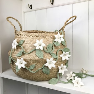 May include: A woven seagrass basket decorated with a crocheted garland of white flowers and green leaves. The garland is draped around the basket and hangs down the side.