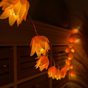 Può includere: Una ghirlanda di luci arancioni a forma di fiore, che emettono un bagliore caldo. Le luci sono infilate insieme, creando una catena decorativa. I fiori sono illuminati dall'interno, fornendo una luce soffusa e invitante.