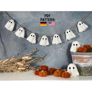Könnte beinhalten: Eine Halloween-Dekoration mit einer Girlande aus weißen Häkelgeistern mit schwarzen Augen, auf einer Schnur aufgereiht. Kleine orangefarbene Häkelkürbisse sind ebenfalls vorhanden. Der Text "PDF PATTERN" ist sichtbar, zusammen mit den Flaggen Deutschlands und der Vereinigten Staaten.