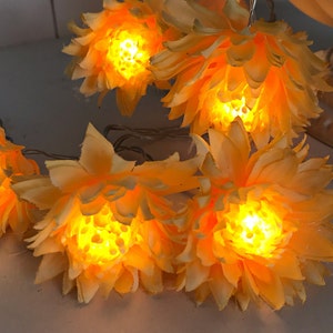 Könnte beinhalten: Eine Lichterkette mit mehreren künstlichen gelben Blumen. Jede Blume hat geschichtete Blütenblätter und ein helles, warmes LED-Licht, das aus der Mitte leuchtet. Die Blumen sind durch einen dünnen, flexiblen Draht miteinander verbunden.