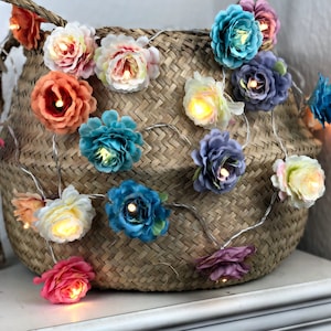 Könnte beinhalten: Eine Lichterkette mit bunten Stoffblumen. Die Lichter sind um einen geflochtenen Korb gewickelt.