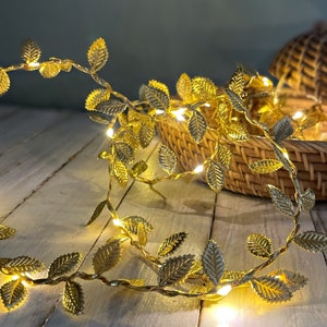 May include: Gold leaf garland with warm white LED lights. The garland is wrapped around a wicker basket.