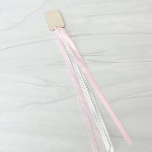 May include: A wooden wand with pink and white ribbons. The ribbons are in varying patterns, including solid pink, white with gold stars, and pink with white polka dots. The wand is rectangular and made of light-coloured wood.