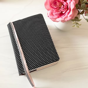 May include: A black notebook with white polka dots and a pink ribbon bookmark. The notebook is closed and has a light pink edge. A vase of pink roses sits in the background.