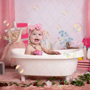May include: A baby girl wearing a pink bow headband sits in a white bathtub with a pink rubber ducky toy. The bathtub is surrounded by pink and green decorations, including a pink curtain, a pink towel, and green leaves.