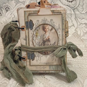 May include: A handmade junk journal featuring a layered cover with vintage-style paper, a central oval cutout displaying a child illustration, the word blessed, a small gold cross charm, metal hinges, and a sage green fabric tie closure.