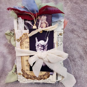 May include: A handmade journal with a vintage aesthetic. It features a collage of images, including an angel statue, sheet music, and decorative frames. The journal is bound with a cream-colored ribbon and adorned with fabric scraps in shades of blue, red, and green.
