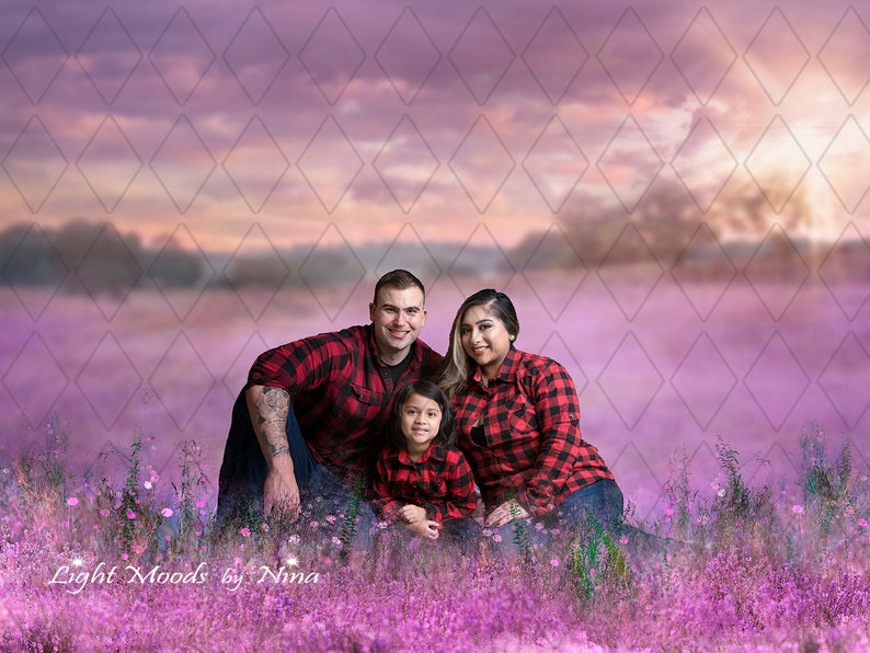 May include: A family of three, a man, a woman, and a child, are sitting in a field of purple flowers. They are all wearing red and black checked shirts. The background is a blurred image of a sunset sky.