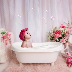 May include: A baby girl wearing a pink bow and a pink bath hat sits in a white bathtub filled with bubbles. The bathtub is on a pink fluffy rug. There are pink flowers and bottles on a shelf in the background.