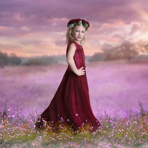 May include: A young girl wearing a red dress and a flower crown stands in a field of purple flowers. The sky behind her is a soft purple and pink.