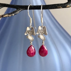 May include: Silver drop earrings featuring a floral design and teardrop-shaped, deep red gemstones. The earrings are suspended from a curved wire and are displayed against a soft blue background.