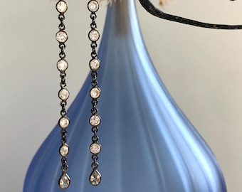 May include: A pair of long, dangling earrings with a dark metal chain and multiple clear, round, sparkling stones. The earrings have a teardrop-shaped stone at the bottom. The background features a blue vase and a dark branch.