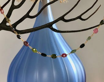 Pode incluir: Um delicado colar de ouro com uma série de pedras preciosas ovais coloridas. As pedras são em tons de rosa, verde e preto. O colar é exibido em um fundo azul suave. As pedras medem aproximadamente 0,5 cm de comprimento.
