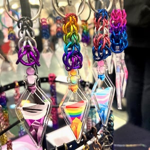 May include: A display of colorful keychains. The keychains feature clear, diamond-shaped pendants with various designs, including a rainbow and a transgender flag. The keychains are attached to metal rings and connected to colorful chainmail. The display is on a black metal rack.