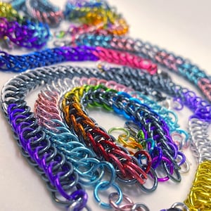 Pode incluir: Uma coleção de joias de cota de malha coloridas, incluindo pulseiras e colares. As peças apresentam uma variedade de tons metálicos, como prata, azul, rosa, roxo e dourado. As malhas são intrincadamente tecidas.