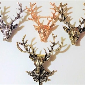 May include: A collection of five metal deer head brooches in various metallic finishes. The brooches feature detailed antlers and are in silver, gold, and copper tones. The deer heads are displayed against a white background.