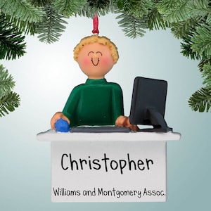 Puede incluir: Un adorno navideño personalizado con un hombre de dibujos animados de pelo rubio sentado en un escritorio con un ordenador. El adorno es blanco con una camisa verde y un ratón azul. El nombre "Christopher" y el texto "Williams and Montgomery Assoc." están impresos en el adorno.