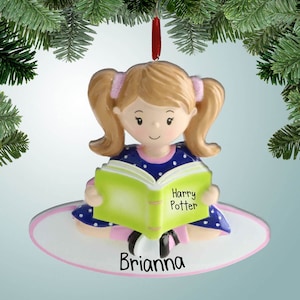 May include: A personalized Christmas ornament featuring a girl with pigtails sitting and reading a green book with the text "Harry Potter" on the cover. The ornament is white with a pink border and the name "Brianna" is written in pink letters.